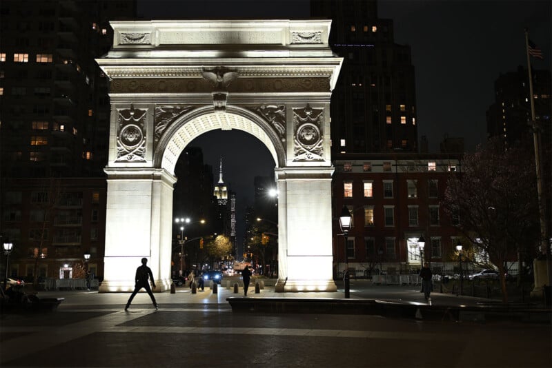 Nighttime view of Washington Square Arch in New York City, illuminated against the dark sky. Silhouetted people walk around the plaza; the Empire State Building is visible in the distance. Trees and surrounding buildings are also dimly lit, adding to the urban atmosphere.