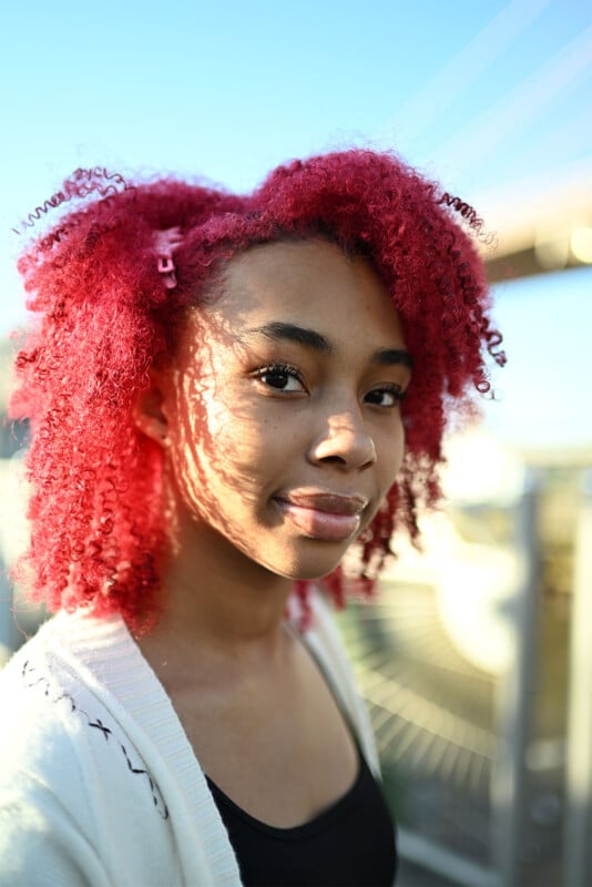 A young person with vibrant, curly pink hair smiles slightly while standing outdoors. They wear a white cardigan over a black top. The background is blurred but shows a bridge structure and blue sky, with sunlight casting shadows on their face.
