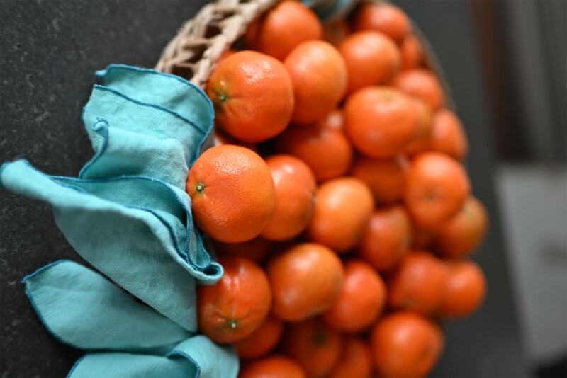 A wicker basket filled with numerous bright orange mandarins is placed on a black surface. A light blue cloth is partially tucked in the basket, draping over the brim and contrasting with the vibrant fruit.