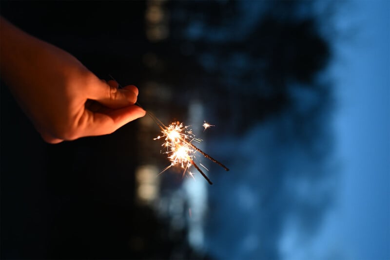 A person holds a lit sparkler against a dark, blurry, outdoor background. The sparkler emits bright, sparkling light, contrasting with the dim surroundings. The image creates a festive, celebratory atmosphere.