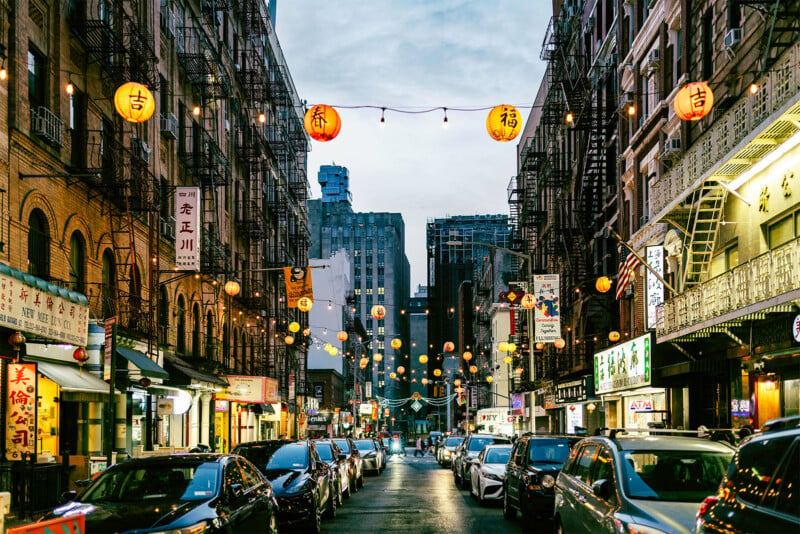 Street scene in a bustling Chinatown at dusk with colorful lanterns hanging overhead. The narrow street is lined with brick buildings, shops, and a variety of parked cars. People are walking on the sidewalks, and lights from the stores add a warm glow to the scene.