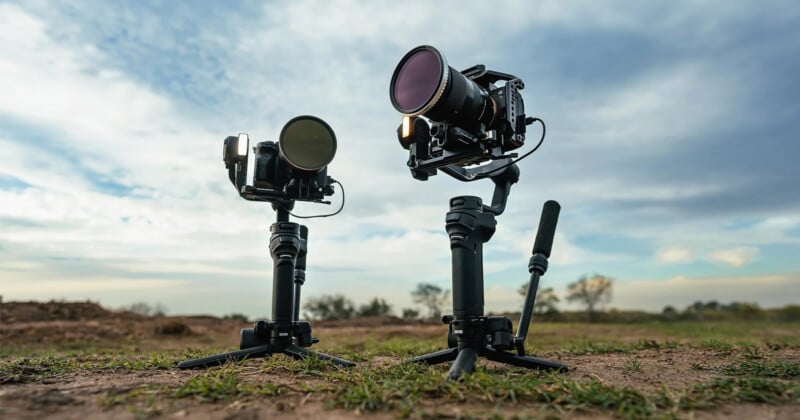 Two professional cameras mounted on mini tripods are set up on a grassy field under a partially cloudy sky. The equipment is positioned at different heights, with the larger camera on the right and the smaller one on the left, capturing the outdoors.
