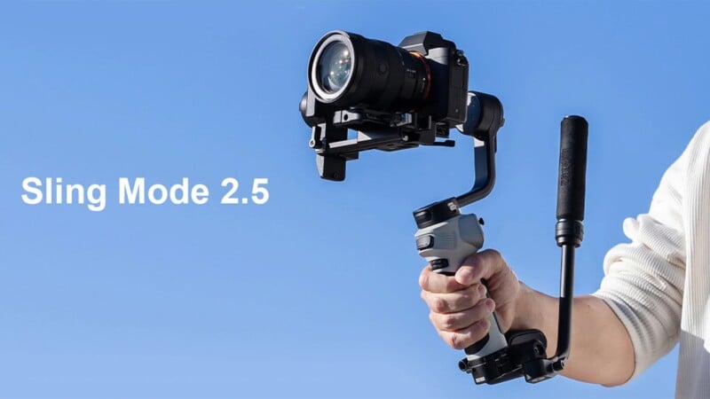 A person holds a camera mounted on a gimbal against a clear blue sky. The text "Sling Mode 2.5" is displayed on the left. The camera is black, and the gimbal has a gray handle with an extended support grip. The person is partially visible, wearing a white shirt.