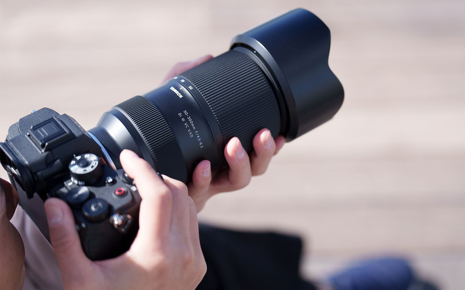 A person holding a professional DSLR camera with a large telephoto lens, adjusting the focus ring. The camera has various dials and buttons visible, and the background is blurred, suggesting an outdoor setting.