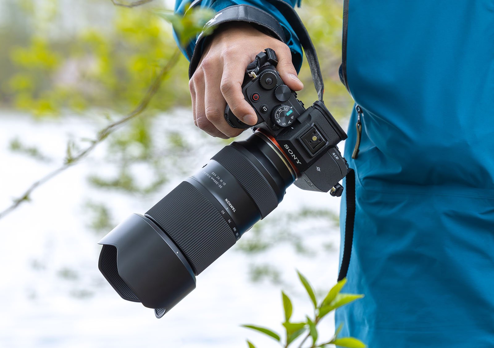 A person in a blue jacket holds a Sony camera with a large telephoto lens attached. The background features blurred greenery and water, indicating an outdoor setting, likely near a lake or river.