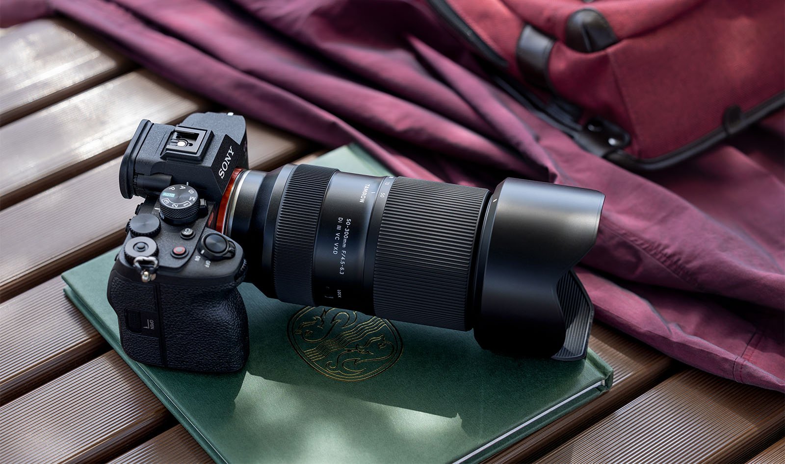 A professional camera with a large lens is placed on a green hardcover book with a crest on the cover. The camera and book rest on a brown wooden surface, and there is a maroon fabric background. A maroon bag partially appears in the background.