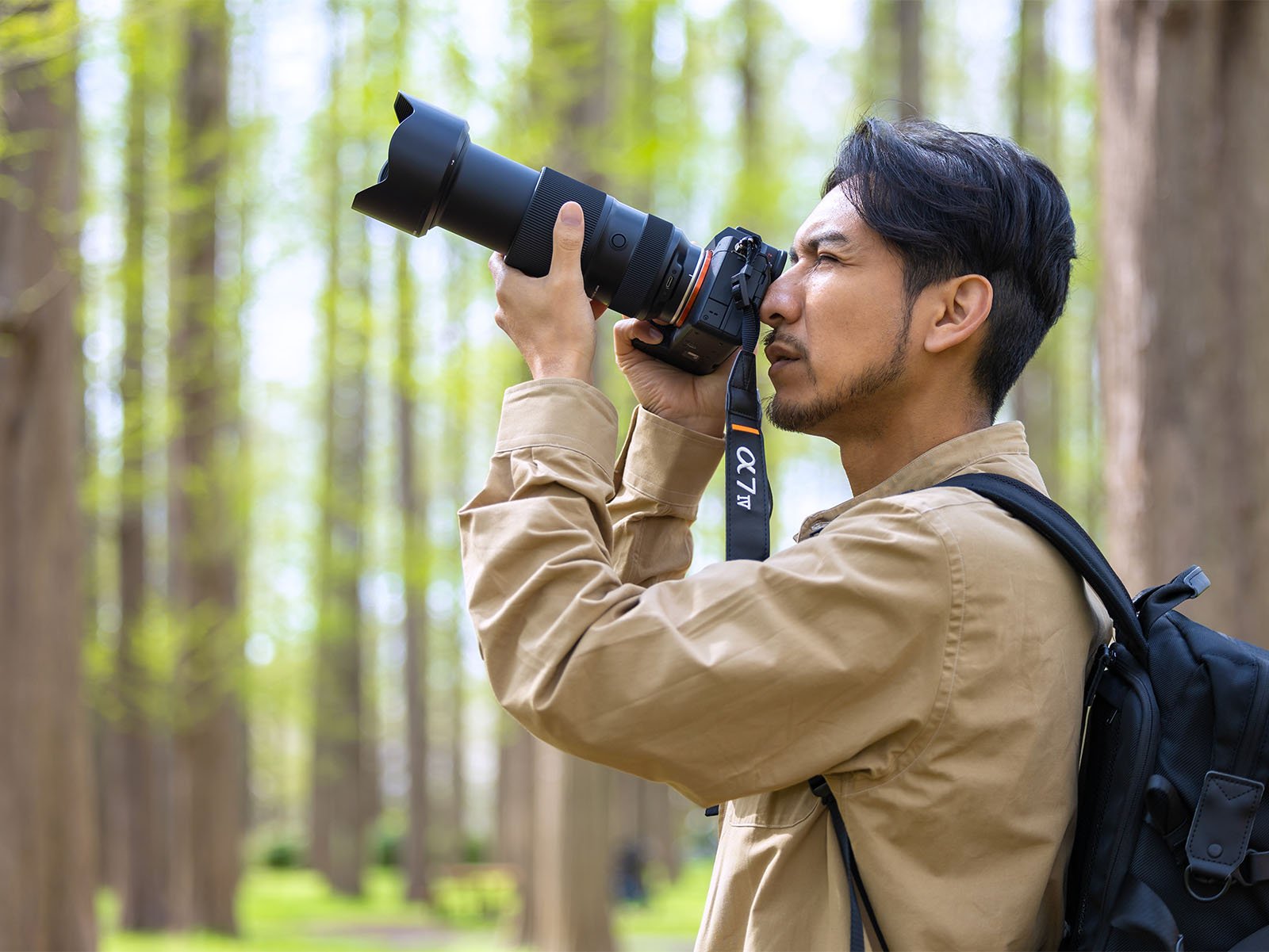 A man wearing a tan jacket holds a large camera with a telephoto lens up to his eye while photographing a forest with tall, leafless trees. He has a black backpack over one shoulder and appears focused on capturing the scene.
