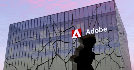 A modern glass building with the Adobe logo prominently displayed at the top right corner. The glass facade of the building appears shattered with large cracks running throughout, creating an illusion of broken glass. The sky in the background has a pinkish hue.