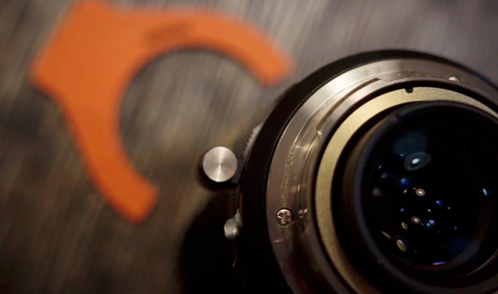 Close-up shot of a camera lens focusing on the rear element and the metal mount. In the blurred background, an orange plastic lens tool is visible. The image highlights details like bolts and glass reflections within the lens.