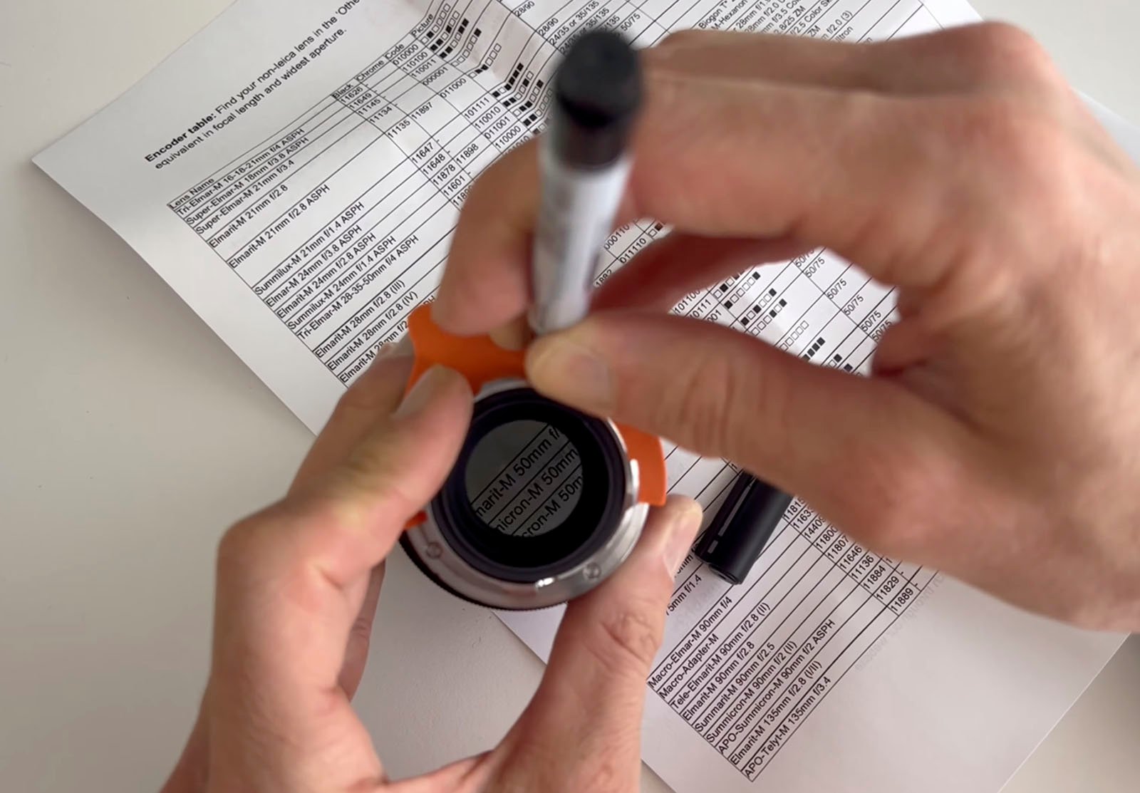 A person is holding an orange and black camera lens adapter and labeling it with a black marker. A printed chart with various details is partially visible on the desk underneath their hands.
