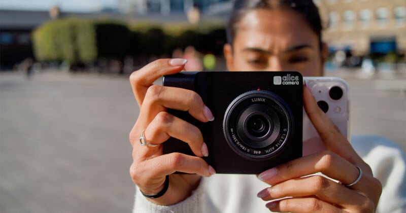 A person holds a smartphone attached to a Lumix camera with their hand, which has rings. The background shows an outdoor setting with blurred trees and buildings, suggesting a sunny day. The person appears to be focusing on capturing an image.