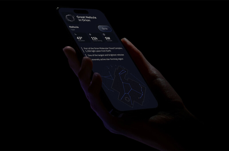 A hand holding a smartphone in the dark displays an astronomy app. The screen shows information about the Great Nebula in Orion, including its magnitude, location details, and a star map of the constellation Orion. The phone is partially illuminated, highlighting the screen.