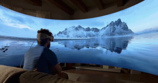 A person wearing a virtual reality headset sits on a couch, immersed in a panoramic scene of snowy mountains and a calm lake that reflects the landscape. The curved display surrounding them enhances the immersive experience.