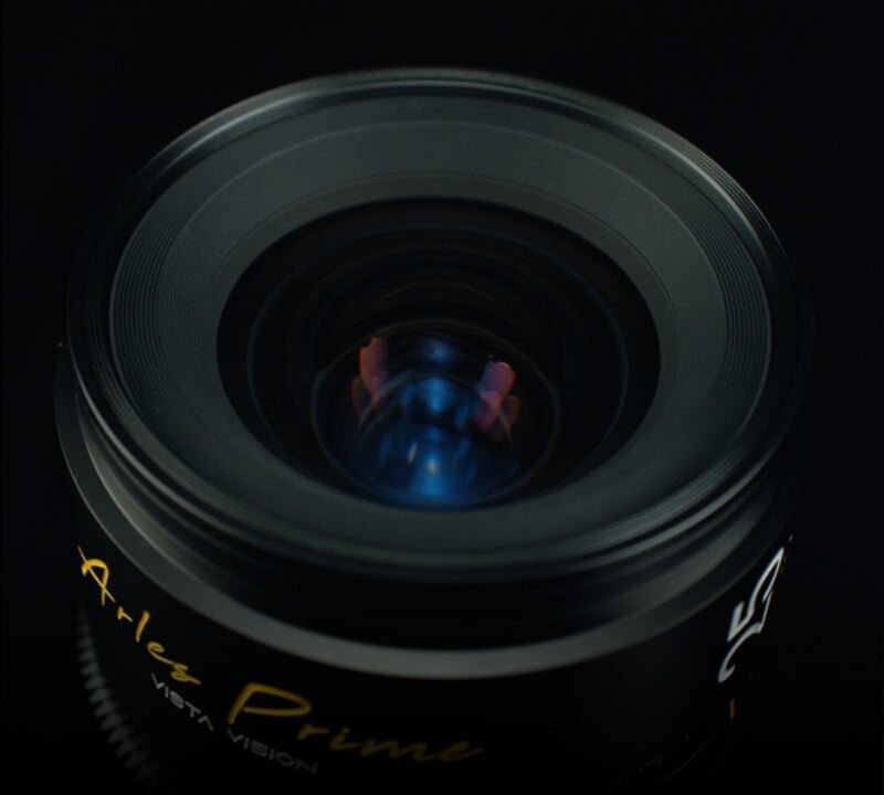 Close-up of a camera lens with the words "Aries Prime" and "Vista Vision" written on it. The outer ring of the lens appears black, and there are reflections of faint blue and red light on the lens surface against a dark background.