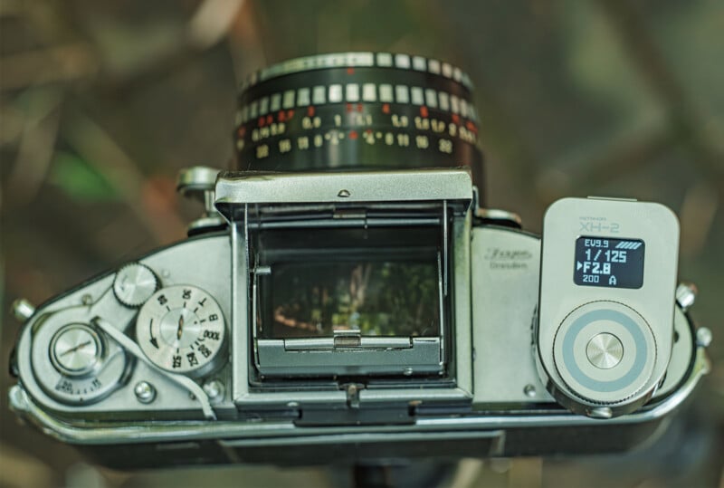 Top view of a vintage film camera with an external digital light meter attached. The camera's viewfinder and various dials are clearly visible, showing aperture, shutter speed, and other settings. The light meter displays exposure information on a small screen.