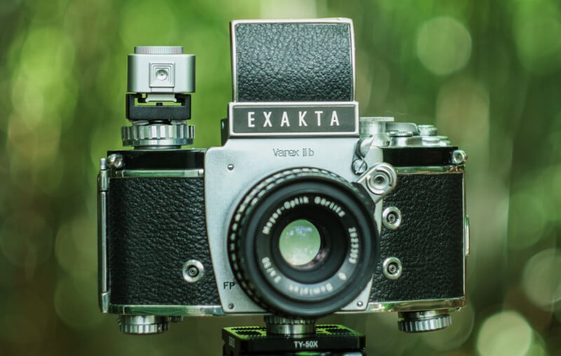 Close-up of an Exakta Varex IIb vintage camera, featuring a black and silver body, mounted on a tripod. The camera is set against a blurred green outdoor background.