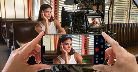 A person in a white tank top with short hair poses inside a diner booth for a video shoot. They rest their head on their hand, looking away. The scene is recorded on a camera, while an individual also views and records it using a smartphone screen in the foreground.