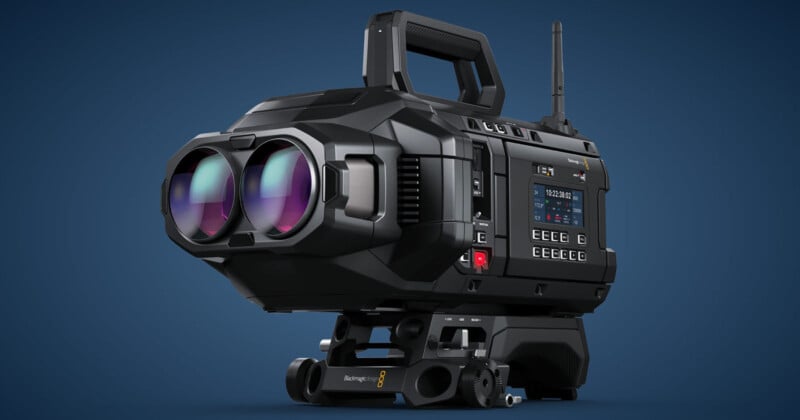 A large black professional video camera with dual lenses, various buttons, and controls. The camera has a handle on top and features a small digital screen displaying various settings and information. The background is a solid blue color.