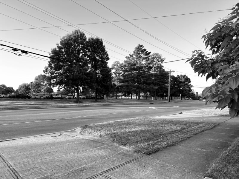 A black-and-white photo of a quiet street scene with intersecting roads, a sidewalk, and overhead power lines. Trees line the far side of the street, and a signpost stands near a traffic light. The image feels peaceful and still.