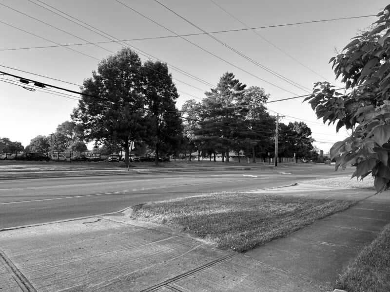 A black-and-white image of an empty street with power lines overhead. Trees line the background, and a well-maintained grassy area is on the right side of the pavement. The scene is calm and devoid of traffic or pedestrians.