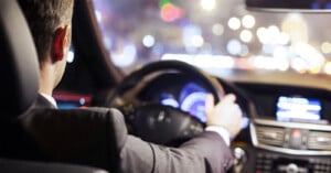 A person wearing a suit is driving a car at night. The image is taken from the backseat, showing the driver's back and hands on the steering wheel. City lights and out-of-focus traffic can be seen through the windshield and side windows.