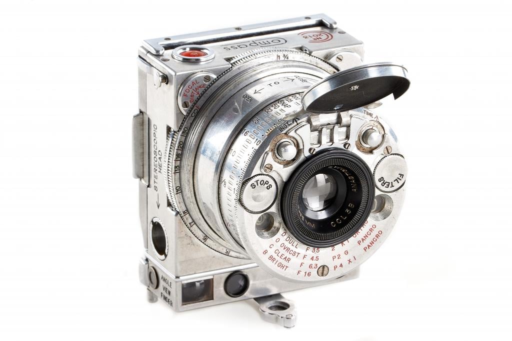 A vintage silver Olympus camera with a collapsible lens and various controls. The lens cap is partially open, revealing the lens elements. The body has multiple dials, buttons, and engraved text in red and black. The camera is set against a white background.