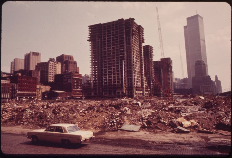 A construction site filled with rubble in an urban area. Partially completed buildings and cranes are visible, with a white car in the foreground. The tallest building in the background is under construction.
