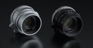 Two camera lenses are shown on a reflective black surface. The lens on the left is silver with "Color-Skopar 50" marked on it, while the lens on the right is black with similar markings. Both lenses feature a sleek, modern design and appear to be of high quality.