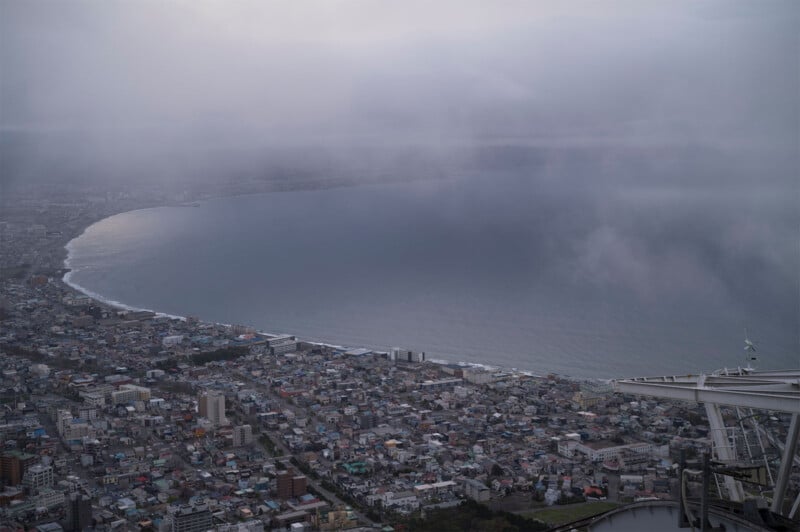 Aerial view of a coastal city with a large bay partially shrouded in dense fog. Buildings densely packed along the coast contrast with the calm, overcast sea. The edge of a tall structure is visible in the foreground.