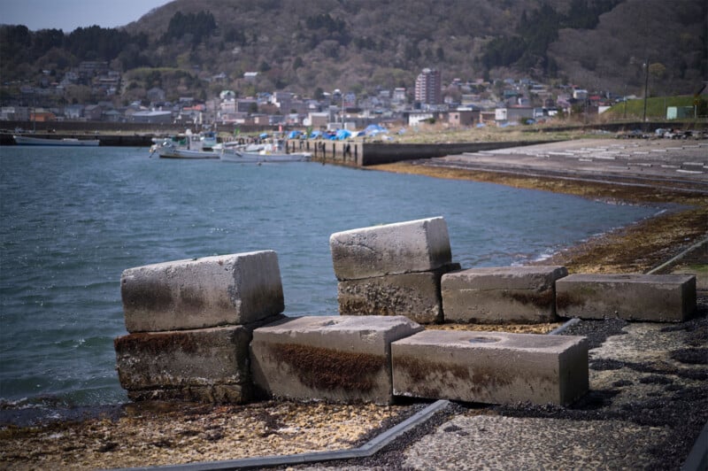Concrete blocks rest along a coast with a calm body of water. In the background, there are several boats docked at the shore and a small town with houses and buildings nestled against a hillside. The sky is clear, and the scene appears peaceful.