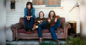 Three men sit on a worn-out couch outside a house. The man on the left has long hair and is wearing a denim jacket, while the middle man holds an acoustic guitar. The man on the right has long hair and a mustache. There's an old barrel and a sign that reads "PARKING" nearby.