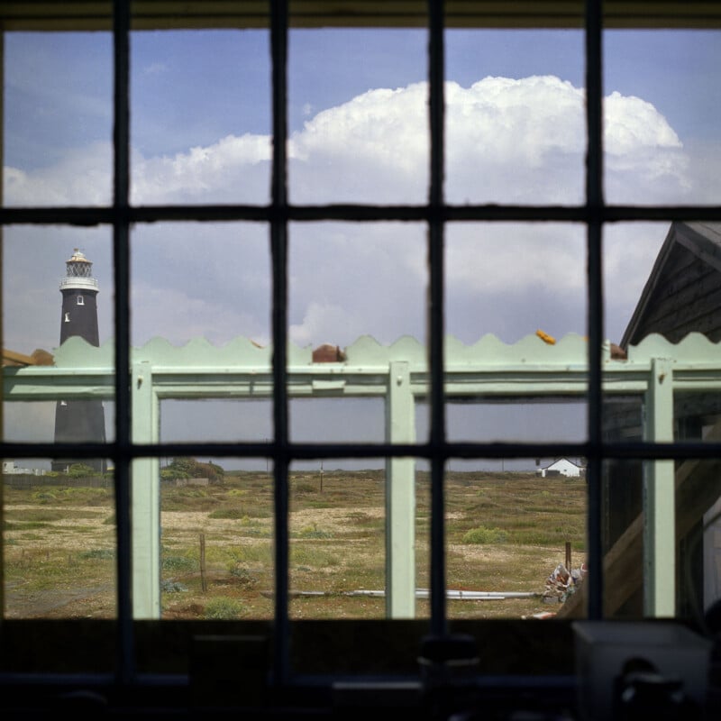 View through a divided window frame looking out at a landscape with a tall, dark lighthouse on the left and a small building in the distance. A garden structure with a scalloped roof edge is visible in the foreground. The sky is partly cloudy.