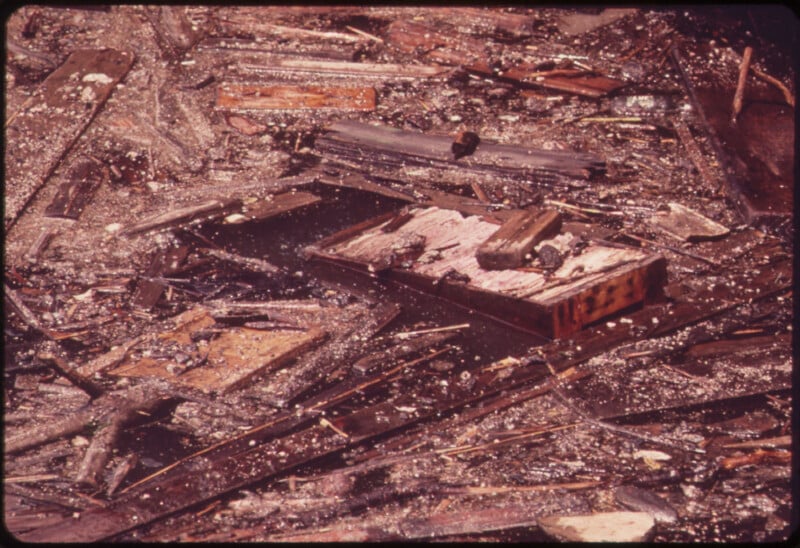 A close-up of scattered debris from a burned building, including charred wooden planks and singed materials. In the center, a partially destroyed briefcase with papers protruding is prominently visible amidst the rubble.