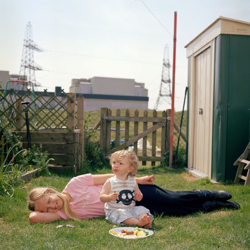 A young child sits on a blanket in a grassy garden holding a plate with food, while an adult with long blonde hair lies nearby, smiling. There is a green shed, wooden fence, and power lines in the background on a sunny day.