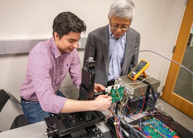 Two individuals are working with electronic equipment. One is adjusting a component on a circuit board, while the other observes. The setup includes wires, connectors, and a multimeter. They are in a laboratory or technical workspace.