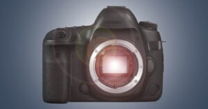 A black DSLR camera without a lens, displaying the exposed image sensor and the lens mount in the center. The camera has a textured grip on the left side, various buttons, and a viewfinder on top. The background is a gradient from dark blue to light gray.