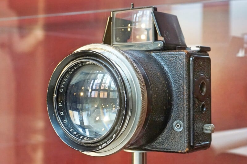 A close-up view of a vintage single-lens reflex (SLR) camera with a large lens, displayed on a stand. The background is blurred, featuring a reddish-brown hue, highlighting the camera's metallic and textured body.