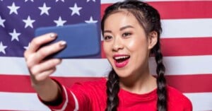 A woman with braided hair smiles while taking a selfie with a blue smartphone. She is wearing a red top and standing in front of an American flag.