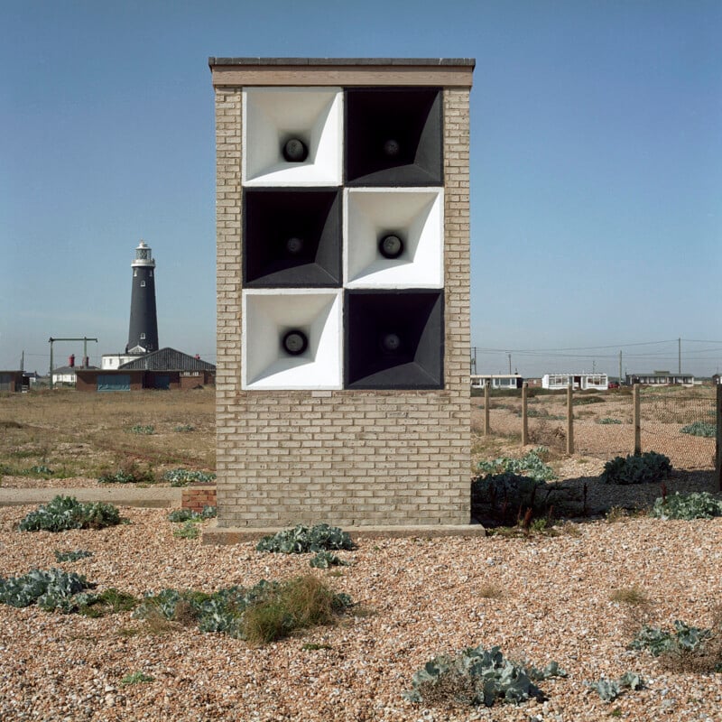 A square brick structure featuring six large speakers is positioned in a gravelly field with sparse vegetation. In the background, a black and white lighthouse stands under a clear blue sky, and small buildings are visible on the horizon.