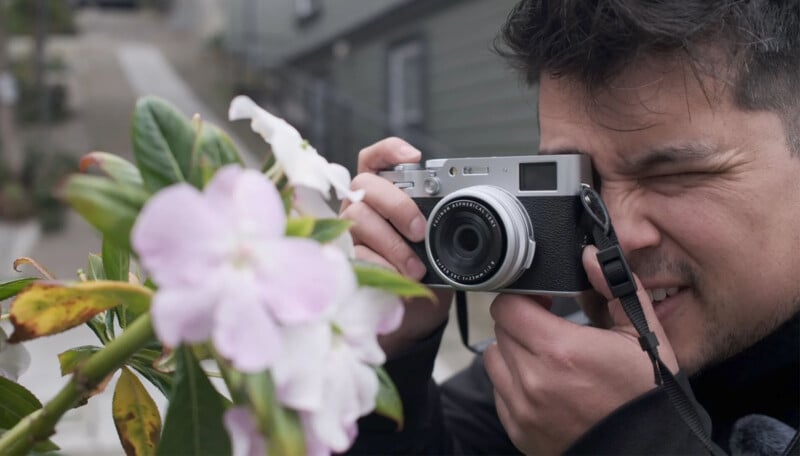 A person with dark hair is using a silver camera to photograph light pink flowers in the foreground. The person is outdoors, and buildings are visible in the background.