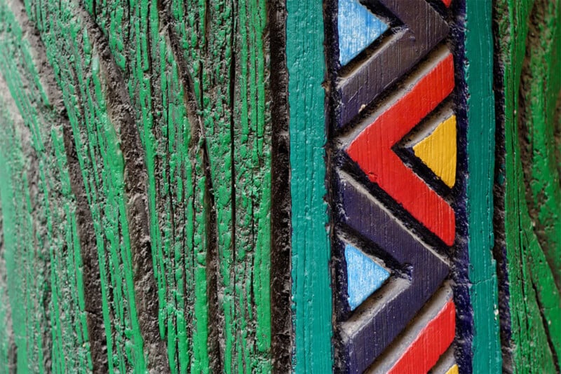 Close-up of a wooden post. The left side is painted green with a rough texture resembling tree bark. The right side features colorful, geometric patterns of triangles painted in red, blue, yellow, and black. The border between the two sections is teal.