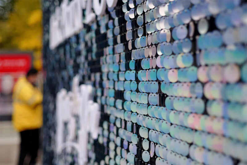 A vibrant wall made of small, reflective, circular discs is in sharp focus on the right side of the image. A person in a yellow jacket is blurred in the background on the left side, standing near a red sign and green foliage.