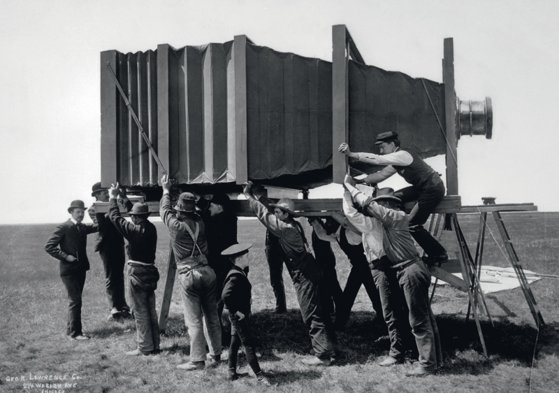 A black-and-white historical photograph shows a group of men assembling or positioning a large, early-era camera on a wooden platform outdoors in a field. The camera is enormous compared to modern standards, emphasizing its unusual size and construction.