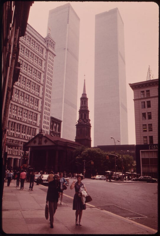 A street scene in New York City featuring the World Trade Center's Twin Towers. The church in the foreground is St. Paul's Chapel. Pedestrians walk along the sidewalk, and people and cars can be seen on the street.