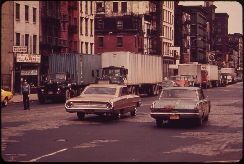 A street scene from the 1970s with several parked trucks and cars. Two vintage cars, one brown and one dark blue, are in the foreground, driving on the cobblestone road. Buildings and a few pedestrians are visible in the background.