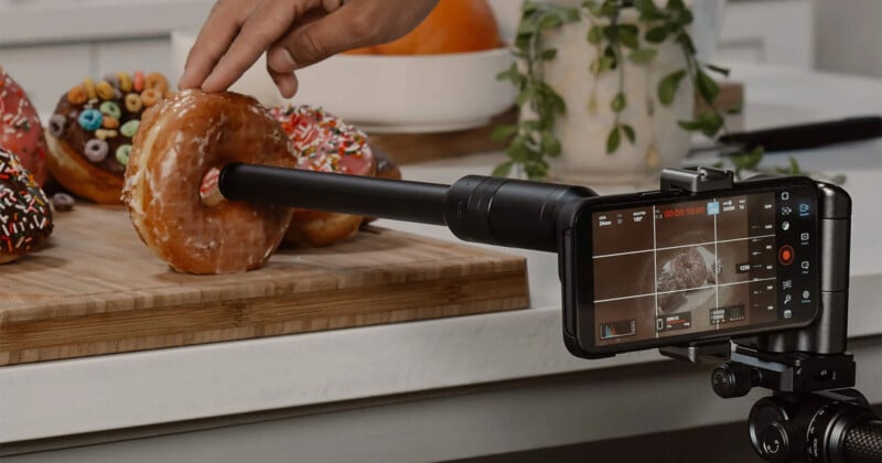 A smartphone mounted on a tripod is being used to take a close-up video of a doughnut on a wooden board. A person's hand is holding the doughnut, and other colorful doughnuts are in the background. A plant is visible in the background as well.
