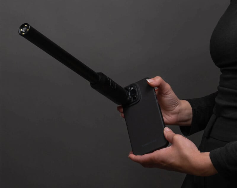A person holding a black gadget that resembles a smartphone attached to a long cylindrical extension, resembling a camera or a detection device, against a dark gray background. The person is wearing a dark, long-sleeve top.
