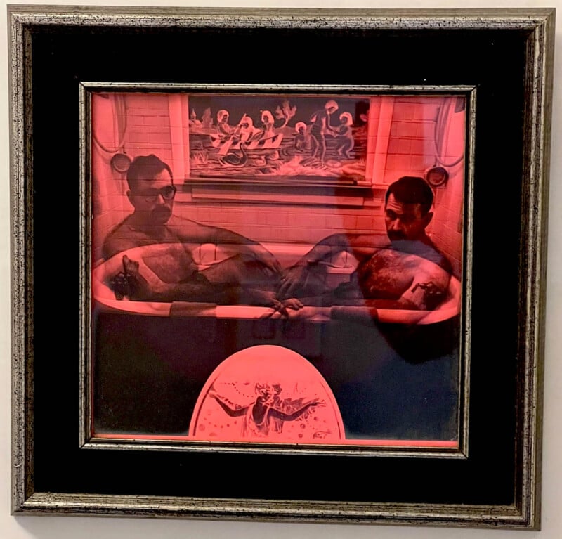 A framed photograph with a red hue depicts two men in a bathtub holding hands. Behind them is a painting showing mythical figures, and a circular emblem with an angelic figure is positioned below.