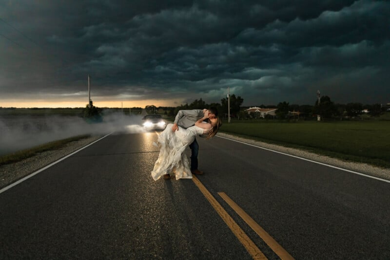 A couple embraces and kisses in the middle of an empty road at dusk, with storm clouds gathering overhead. The groom dips the bride, whose dress touches the pavement. A car approaches with headlights on, adding a dramatic effect to the scene.