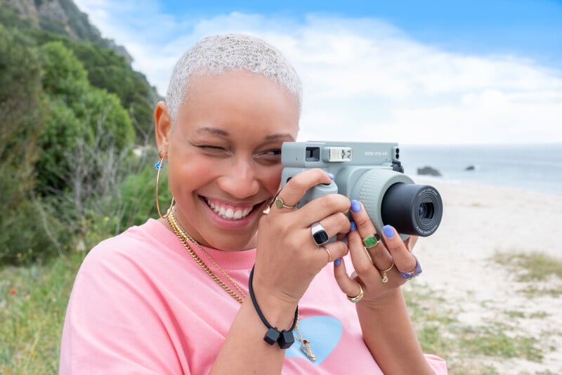 A person with short, blond hair is outdoors, holding a camera to their eye and smiling. They are wearing a light pink shirt and several rings. The background features a beach with sand, greenery, and a cloudy blue sky.